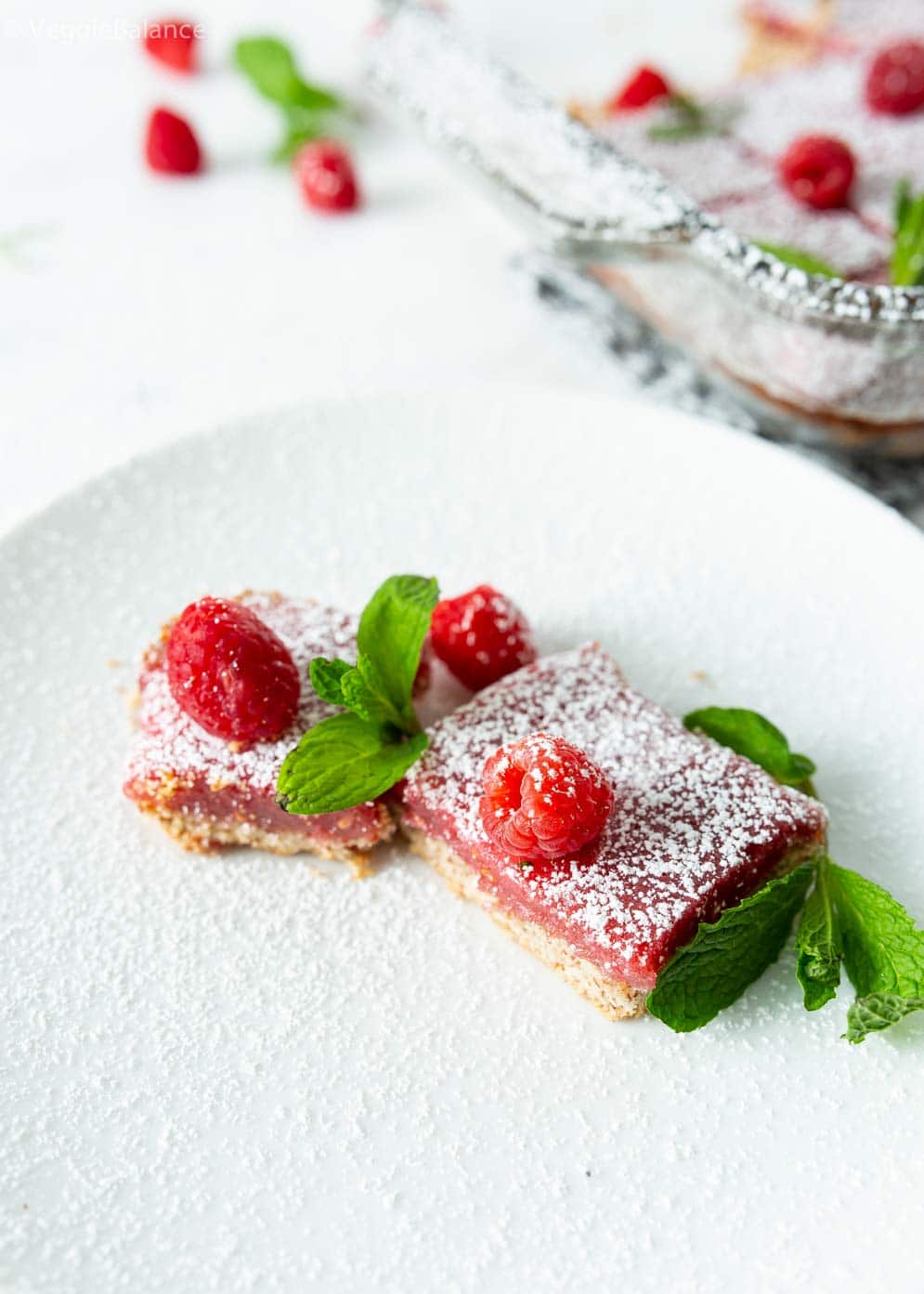 Two Vegan Raspberry Lemon Bars with fresh raspberries and mint leaves decorated around on a small plate